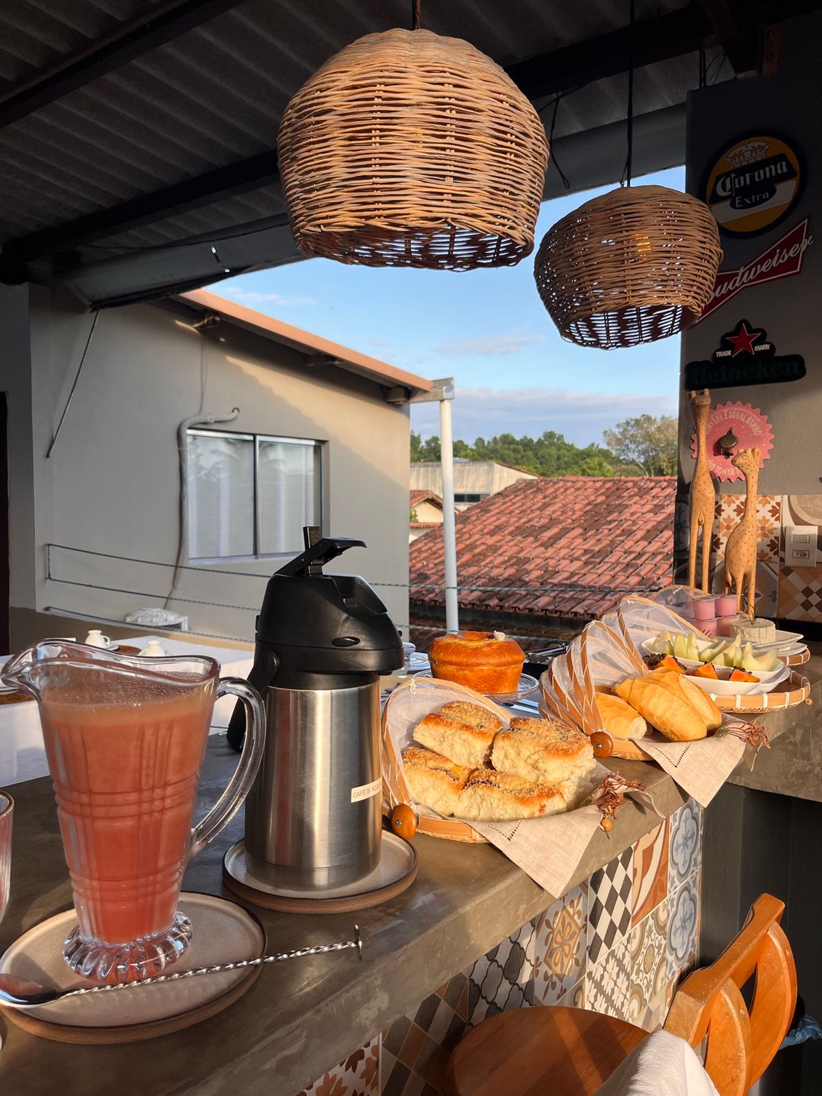 Mesa do café com suco natural, pães e bolos, com vista para o telhado ao fundo